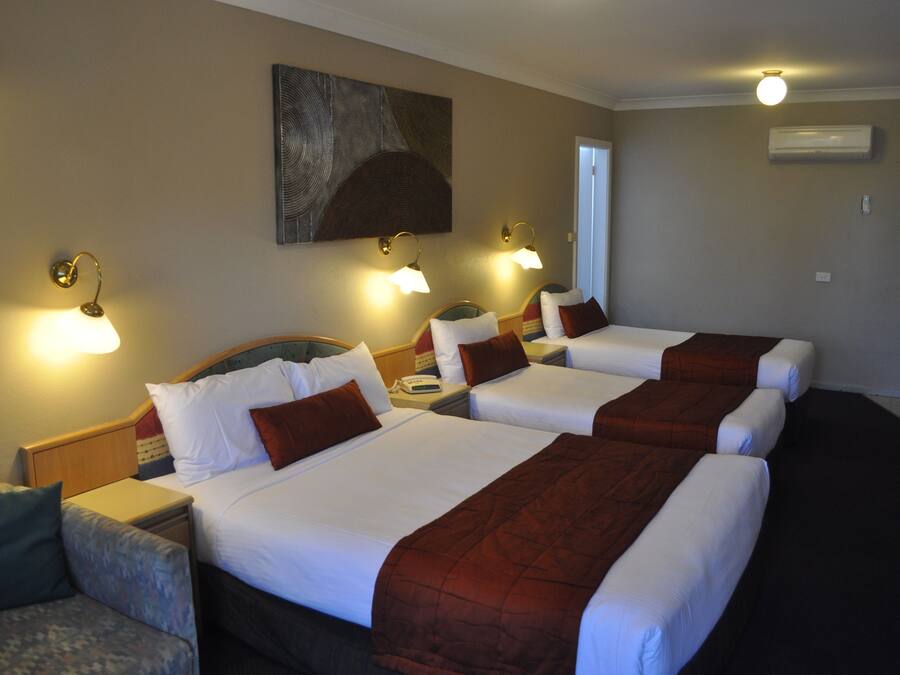 Habitación de hotel con tres camas, cada una con ropa blanca y cabeceras marrones. Dos lámparas de pared a cada lado de las camas.