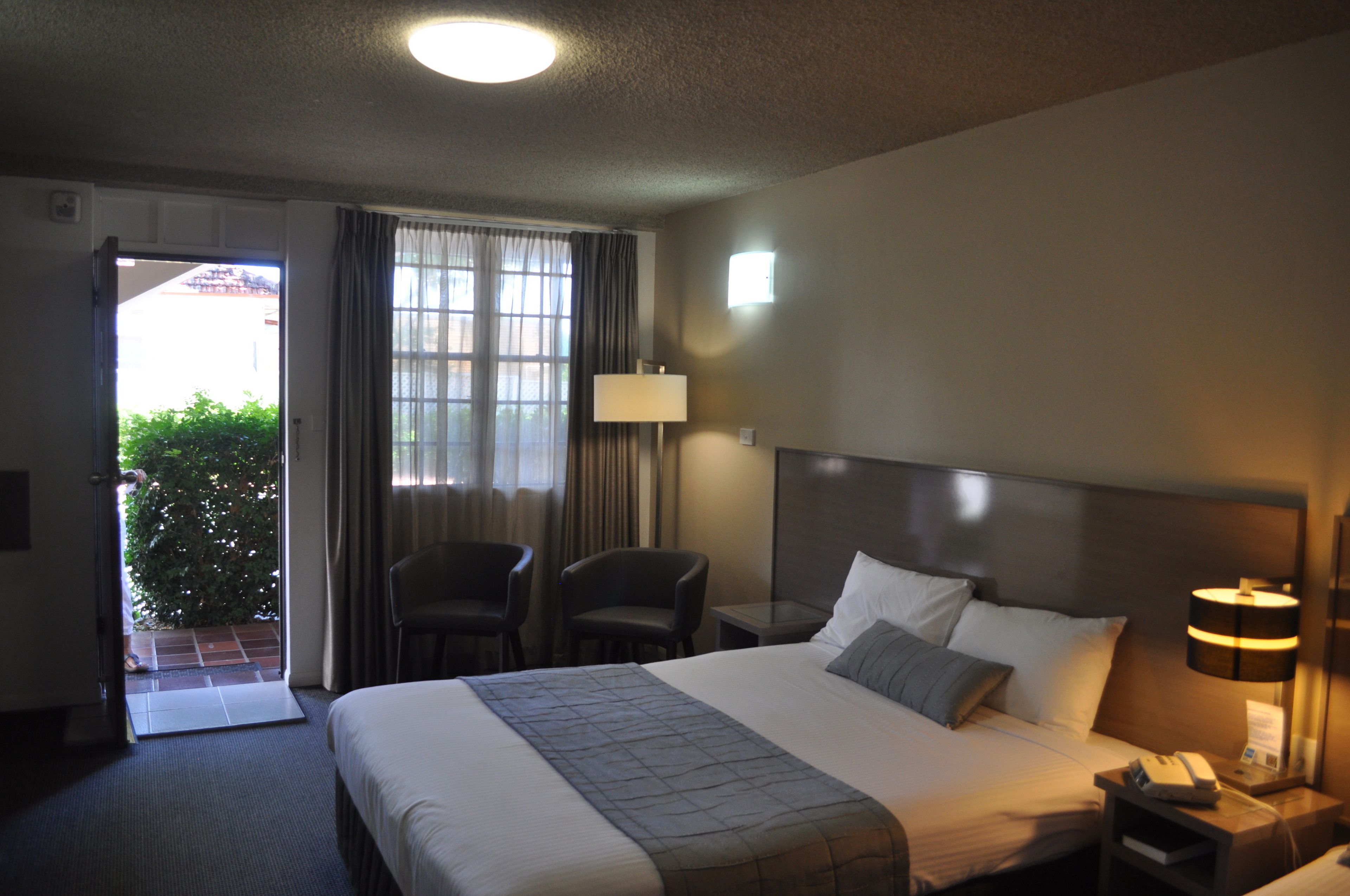 A hotel room with a bed, a nightstand, a lamp, a chair, and a view of a green plant outside.