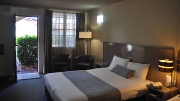 A hotel room with a bed, a nightstand, a lamp, a chair, and a view of a green plant outside.