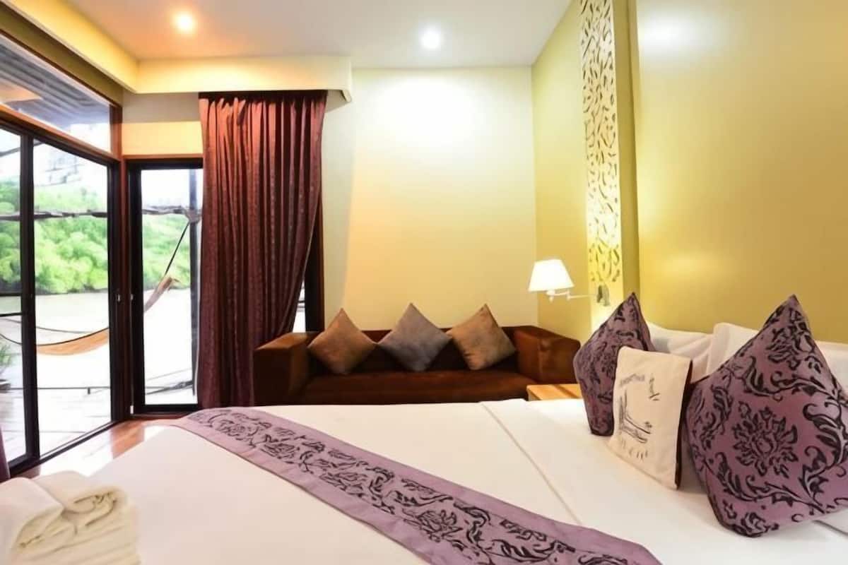 A hotel room with a bed, a sofa, a lamp, and a view of greenery through the window.