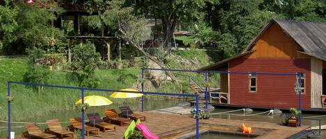 Un muelle de madera con sillas de descanso y sombrillas, un pequeño edificio y una masa de agua.