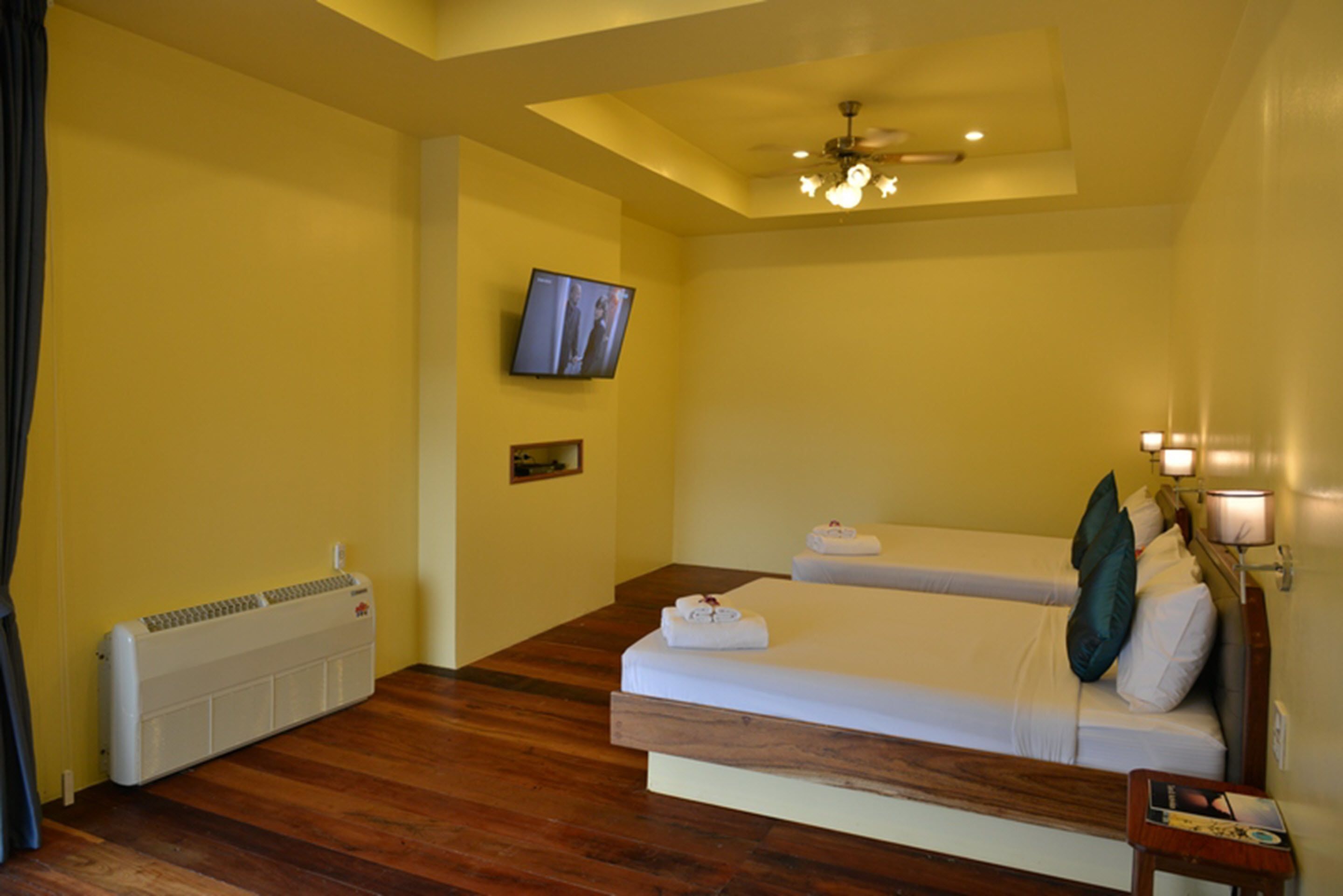 Une chambre d’hôtel avec un plancher en bois, deux lits simples, un ventilateur de plafond, un téléviseur fixé au mur et un radiateur.