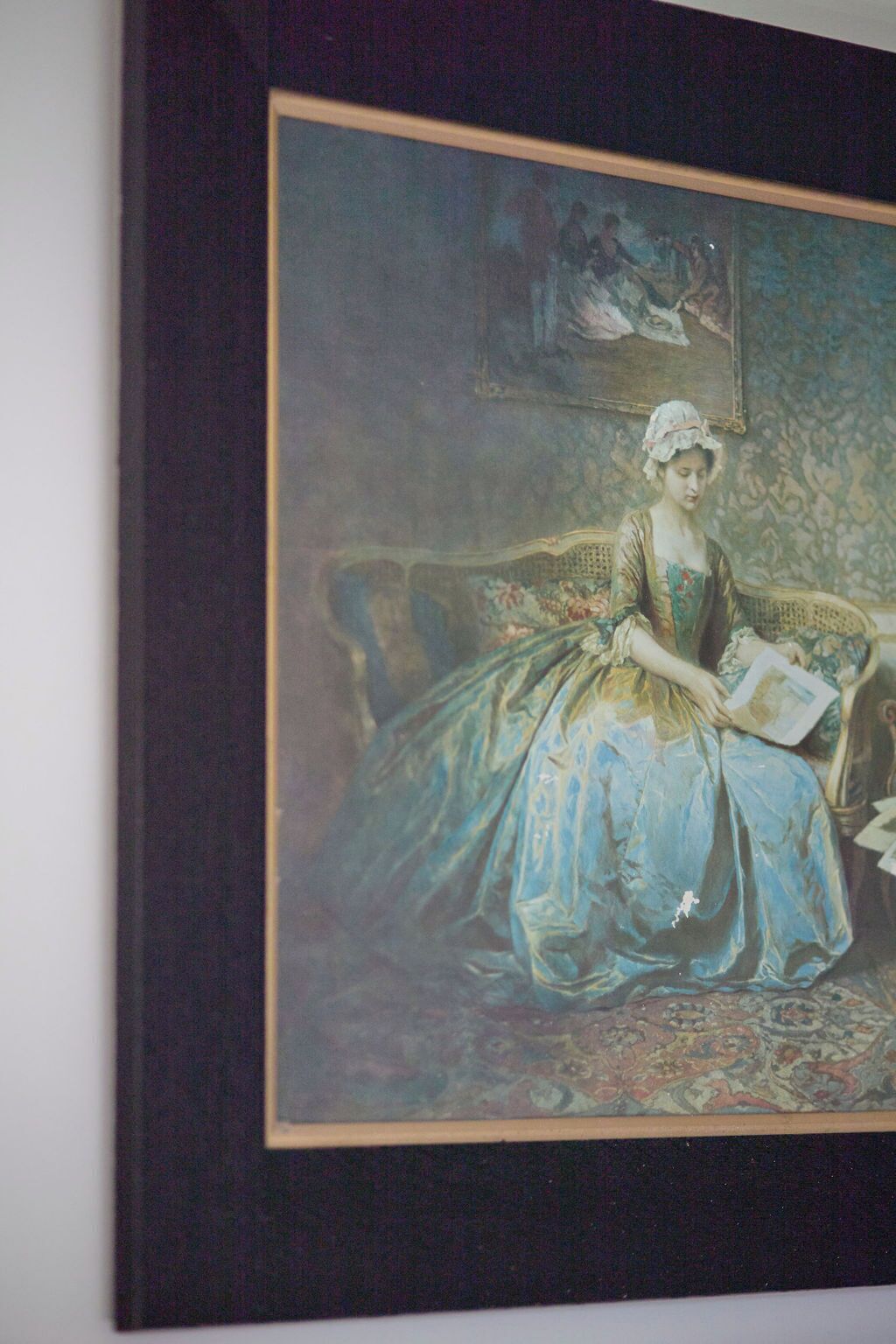 Ein gerahmtes Gemälde, das eine Frau in einem blauen Kleid sitzend auf einem Sofa mit einem gemusterten Teppich zeigt.