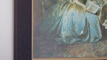 Ein gerahmtes Gemälde, das eine Frau in einem blauen Kleid sitzend auf einem Sofa mit einem gemusterten Teppich zeigt.