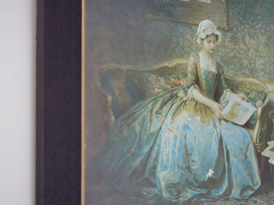 Ein gerahmtes Gemälde, das eine Frau in einem blauen Kleid sitzend auf einem Sofa mit einem gemusterten Teppich zeigt.