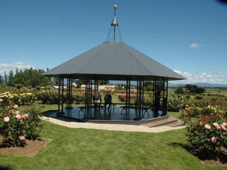 A gazebo with a metal roof and a central pole, surrounded by well-maintained flower beds and a grassy lawn.
