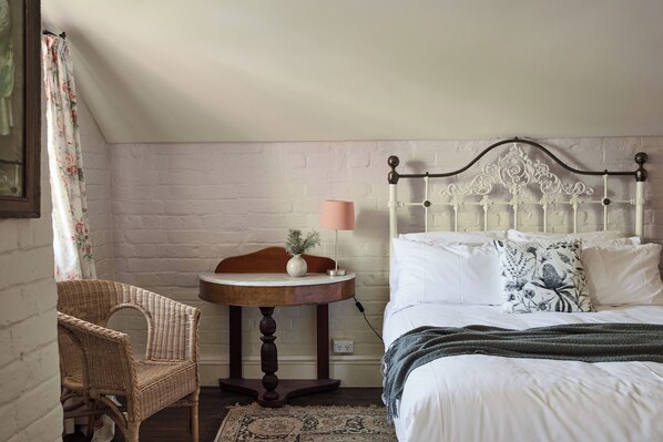 A bedroom with a bed, a round wooden table with a lamp, a wicker chair, and a white brick wall.