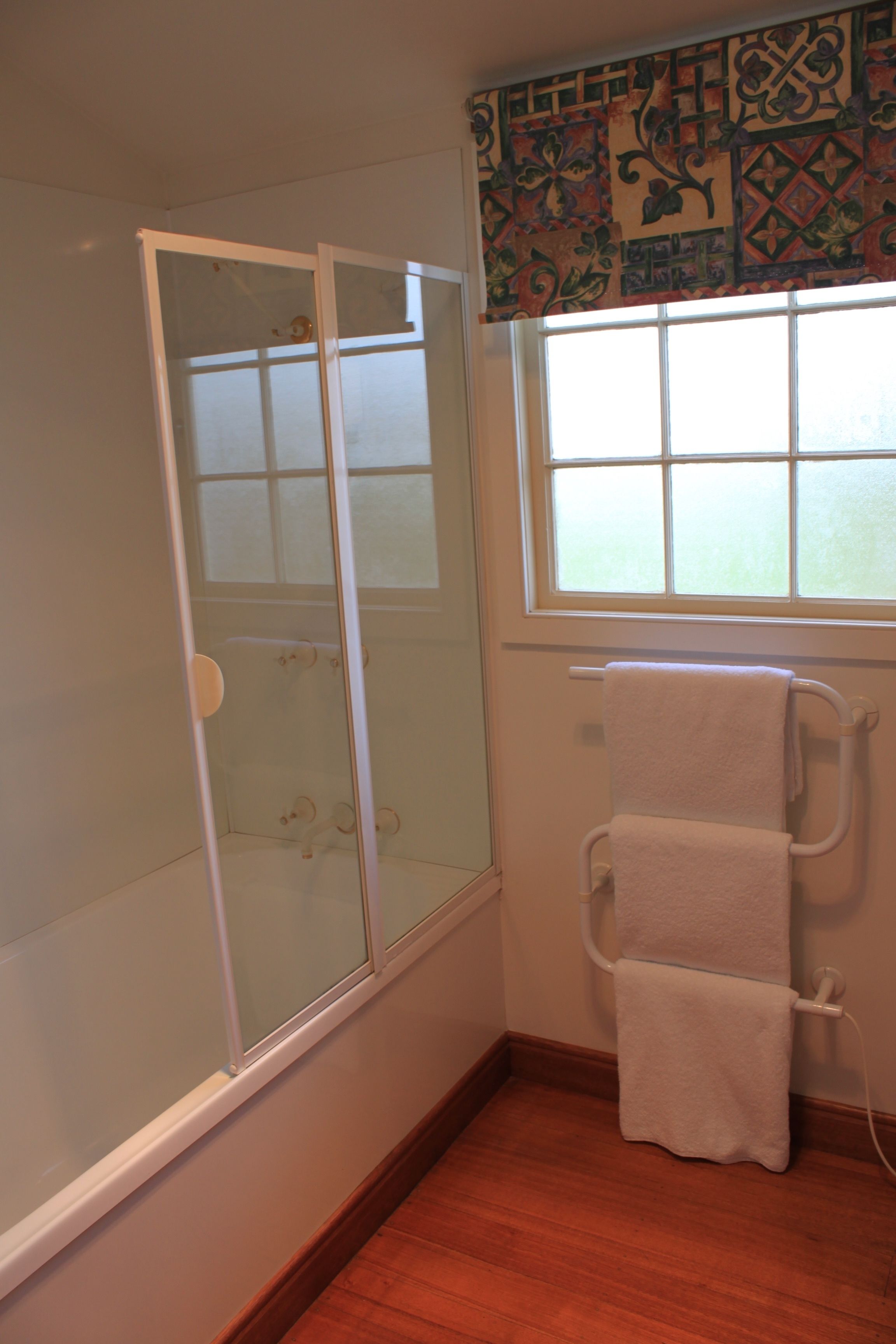 A bathroom with a glass shower enclosure, a window with patterned curtains, and a towel rack with multiple towels.