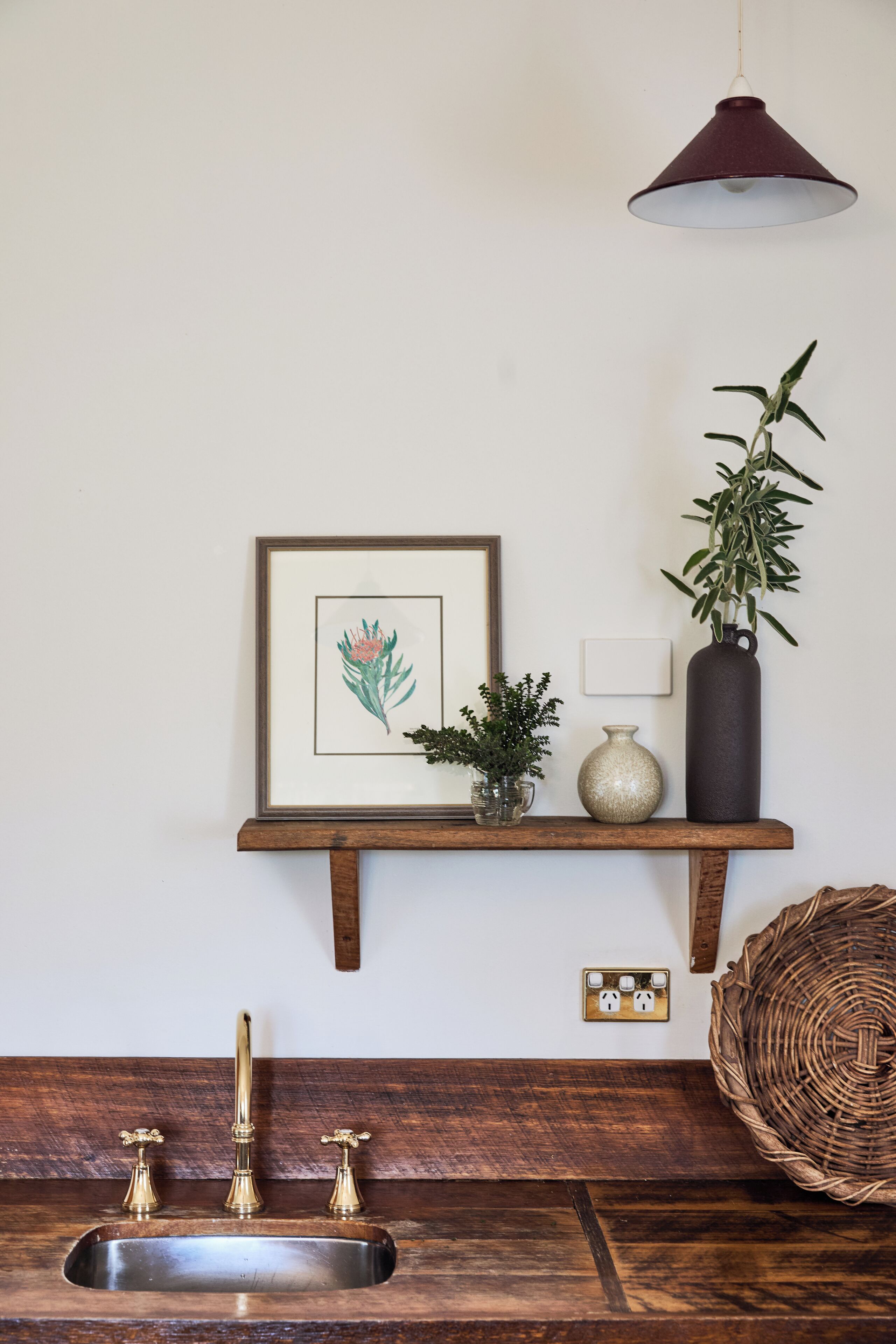 A kitchen with a wooden countertop, a sink with three faucets, a framed botanical print, a potted plant, a vase, and a hanging light fixture.