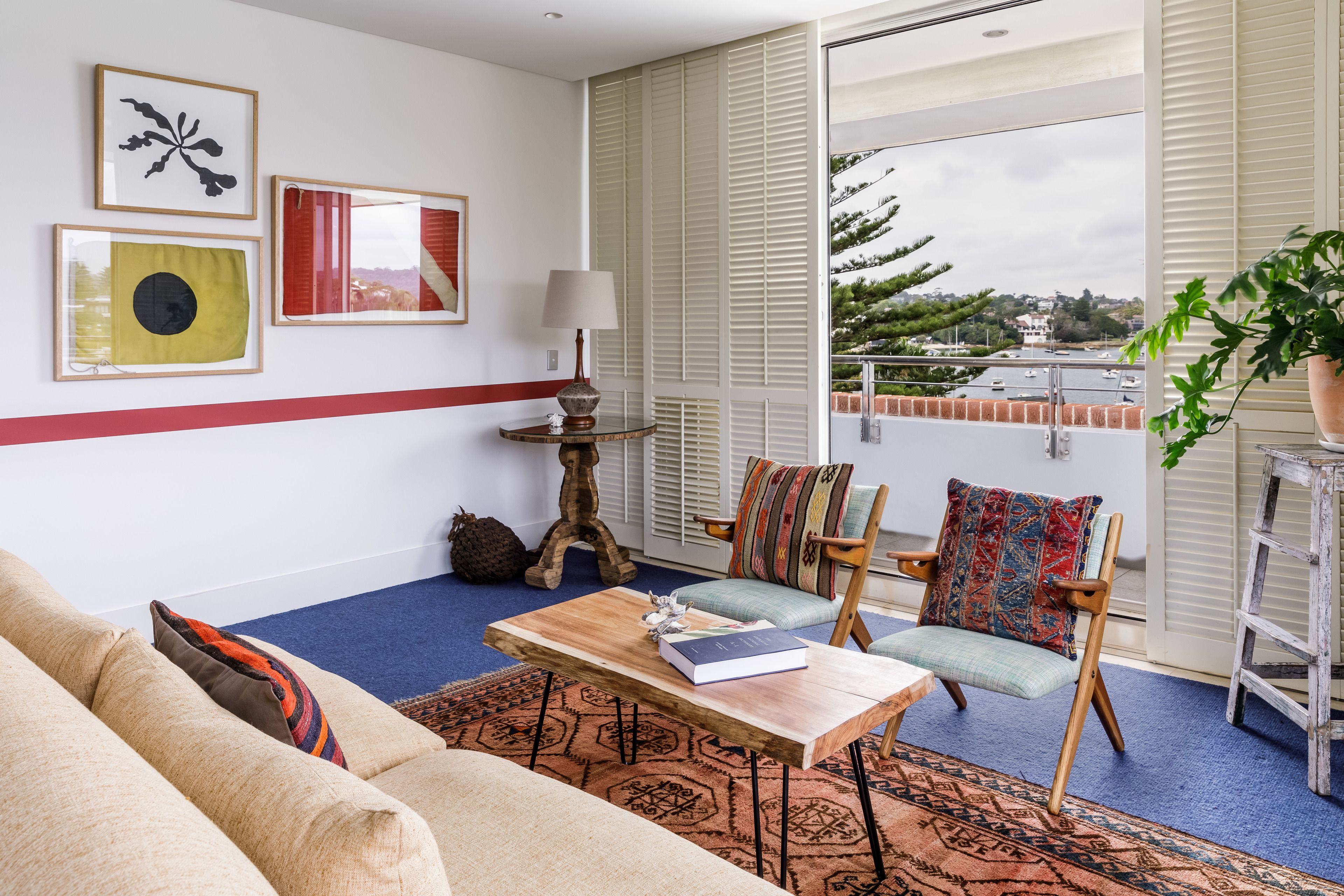 A living room with a sofa, a coffee table with a book, a chair with a colorful cushion, a lamp, and a view of the outdoors through large windows.