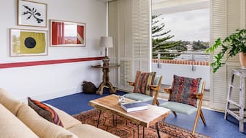 A living room with a sofa, a coffee table with a book, a chair with a colorful cushion, a lamp, and a view of the outdoors through large windows.