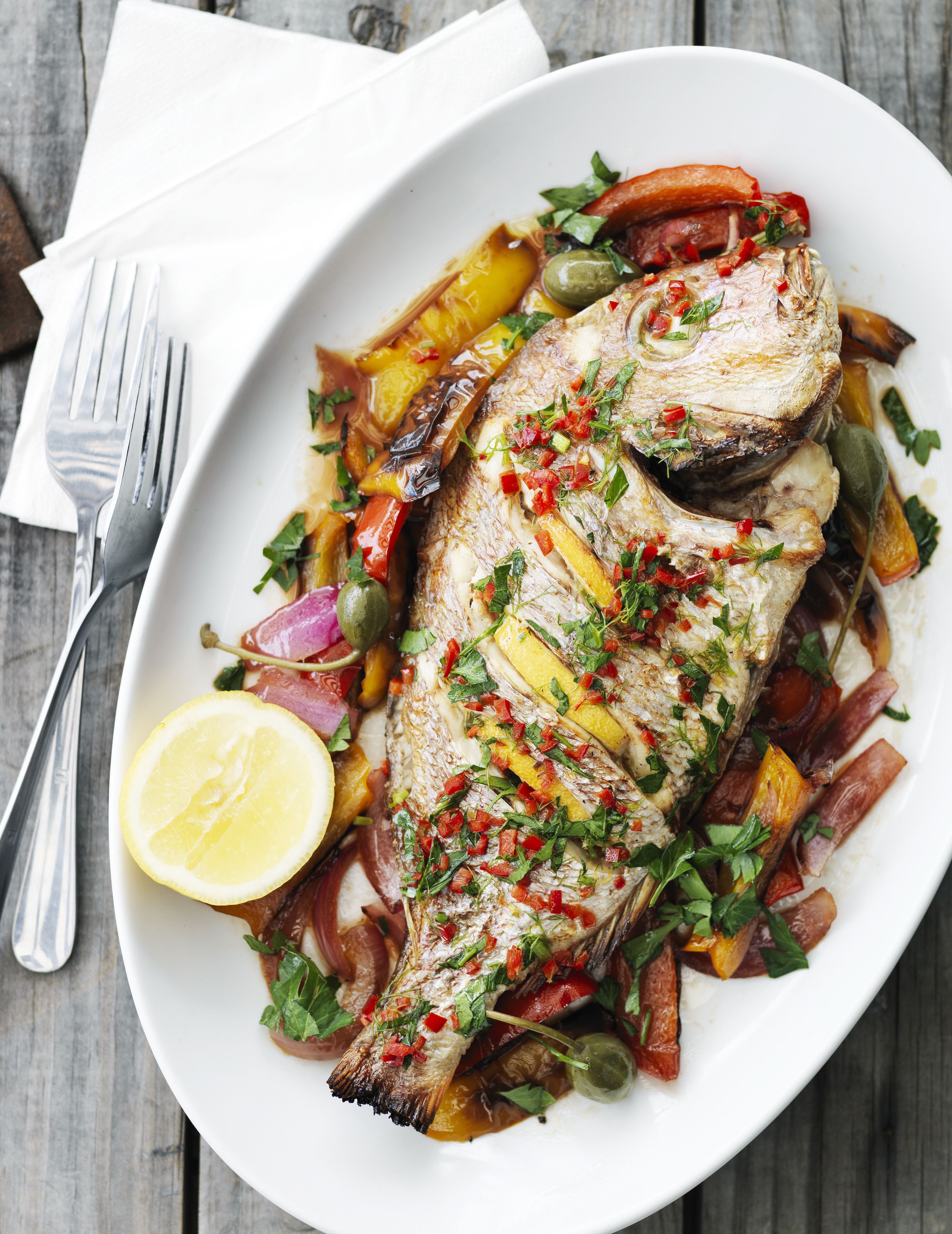 A white oval plate with a whole fish garnished with herbs and vegetables, accompanied by a lemon wedge and a fork on a wooden table.
