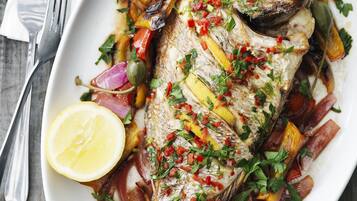 A white oval plate with a whole fish garnished with herbs and vegetables, accompanied by a lemon wedge and a fork on a wooden table.