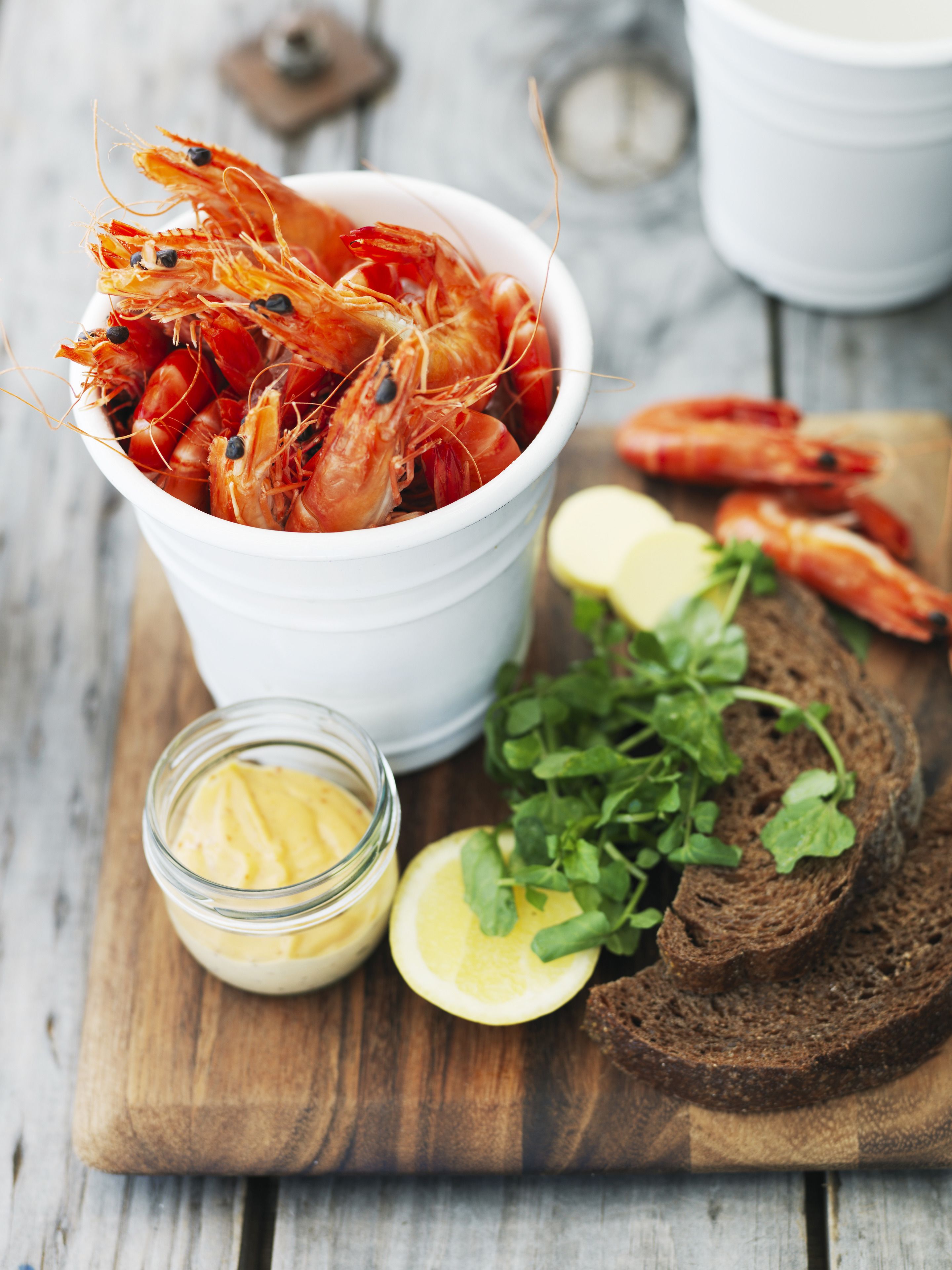 A wooden cutting board with a bowl of cooked shrimp, a jar of butter, lemon slices, and a slice of dark bread.