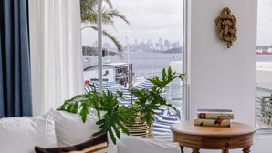 Une pièce avec un canapé blanc, un coussin à rayures, une table ronde en bois et une vue sur les toits d’une ville et sur l’eau, le tout visible à travers une grande fenêtre.