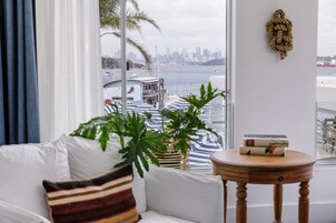 Une pièce avec un canapé blanc, un coussin à rayures, une table ronde en bois et une vue sur les toits d’une ville et sur l’eau, le tout visible à travers une grande fenêtre.