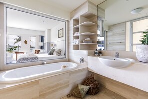 A modern bathroom with a large mirror, two sinks, and a bathtub. There are shelves above the sinks and a potted plant on the right.