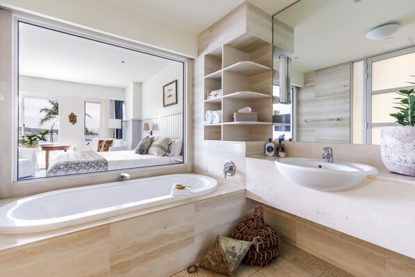 A modern bathroom with a large mirror, two sinks, and a bathtub. There are shelves above the sinks and a potted plant on the right.