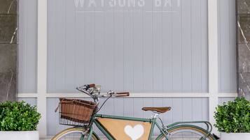 A green bicycle with a wicker basket and a brown seat parked in front of a grey door with the text "Watsons Bay" and "5JOTIQUE HOTEL" on it.