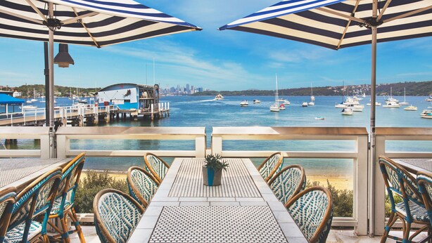 Une terrasse agrémentée de tables et de chaises, d’où l’on peut admirer un port de plaisance avec ses bateaux, ainsi qu’un panorama sur la ville au loin.