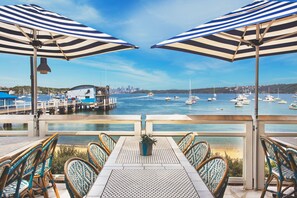 A terrace with tables and chairs, overlooking a marina with boats and a city skyline in the distance.
