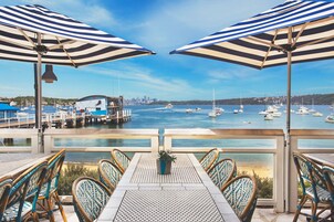 Une terrasse agrémentée de tables et de chaises, d’où l’on peut admirer un port de plaisance avec ses bateaux, ainsi qu’un panorama sur la ville au loin.