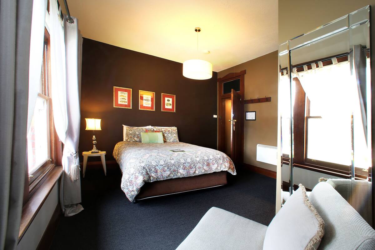 A hotel room with a bed, a bedside table with a lamp, a large mirror, and framed pictures on the wall.