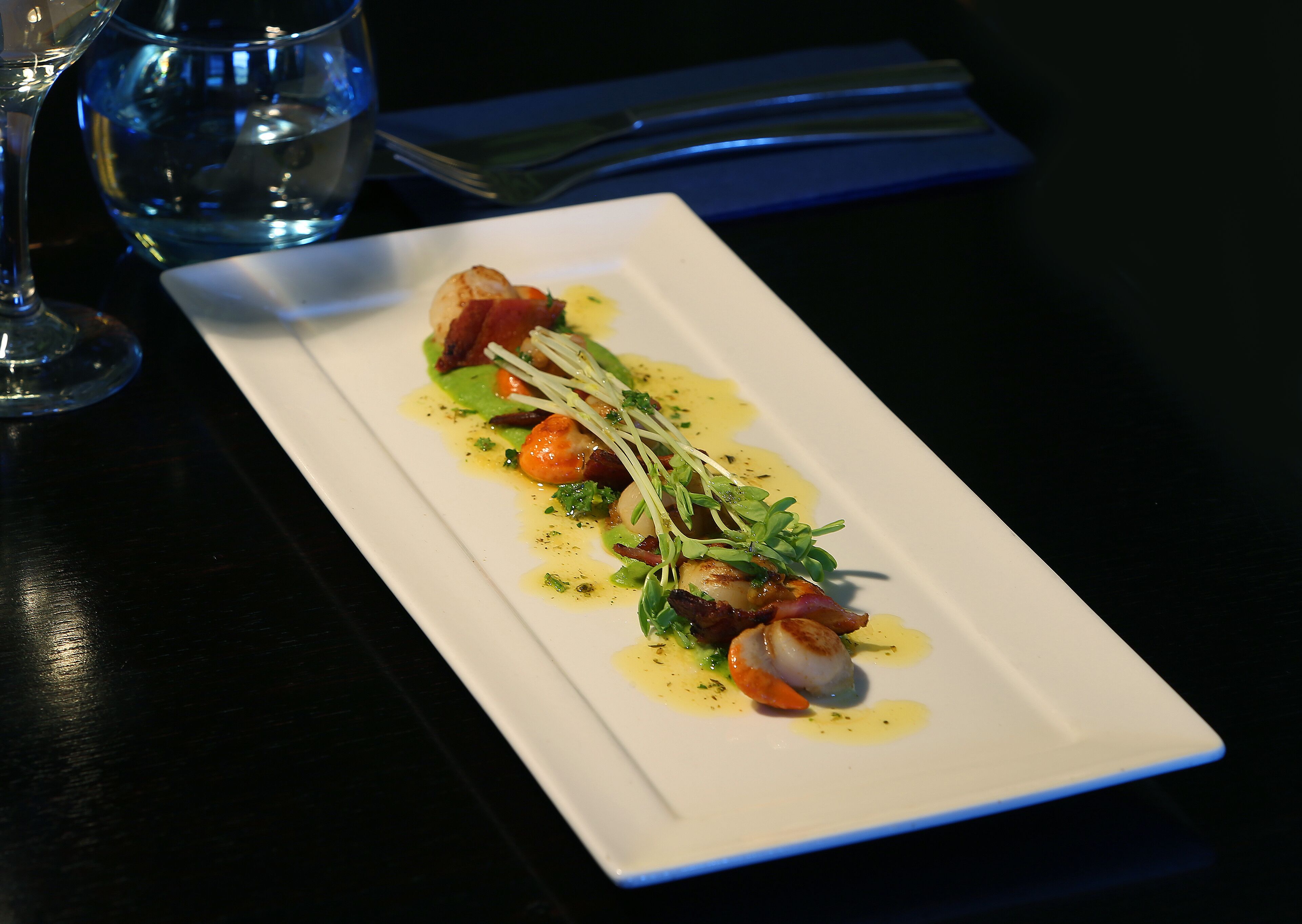Un plato rectangular blanco con un plato de mariscos, adornado con hierbas y un chorrito de salsa, sobre una mesa oscura.