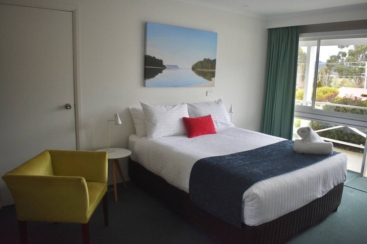 A hotel room with a bed, a yellow chair, a small table, and a painting of a lake.