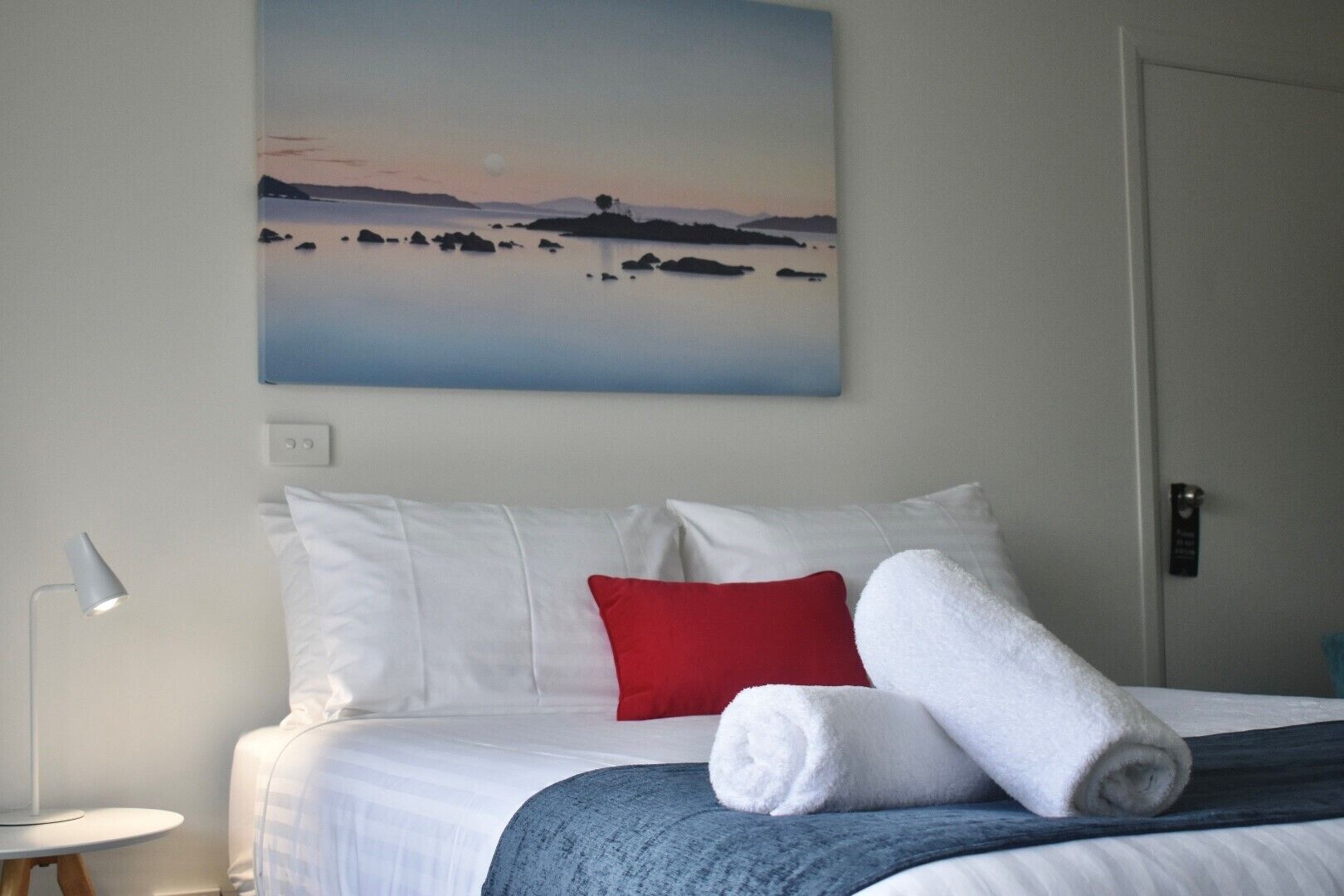 A hotel room with a bed, white and red pillows, a rolled white towel, a blue blanket, a painting of a sunset over water, a bedside lamp, and a door with a sign.