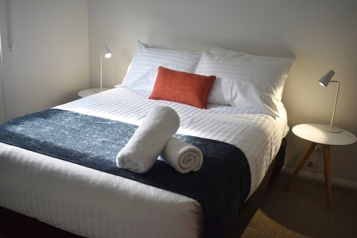 A neatly made bed with white and blue bedding, a rolled white towel, and an orange pillow. There are two bedside tables with lamps.
