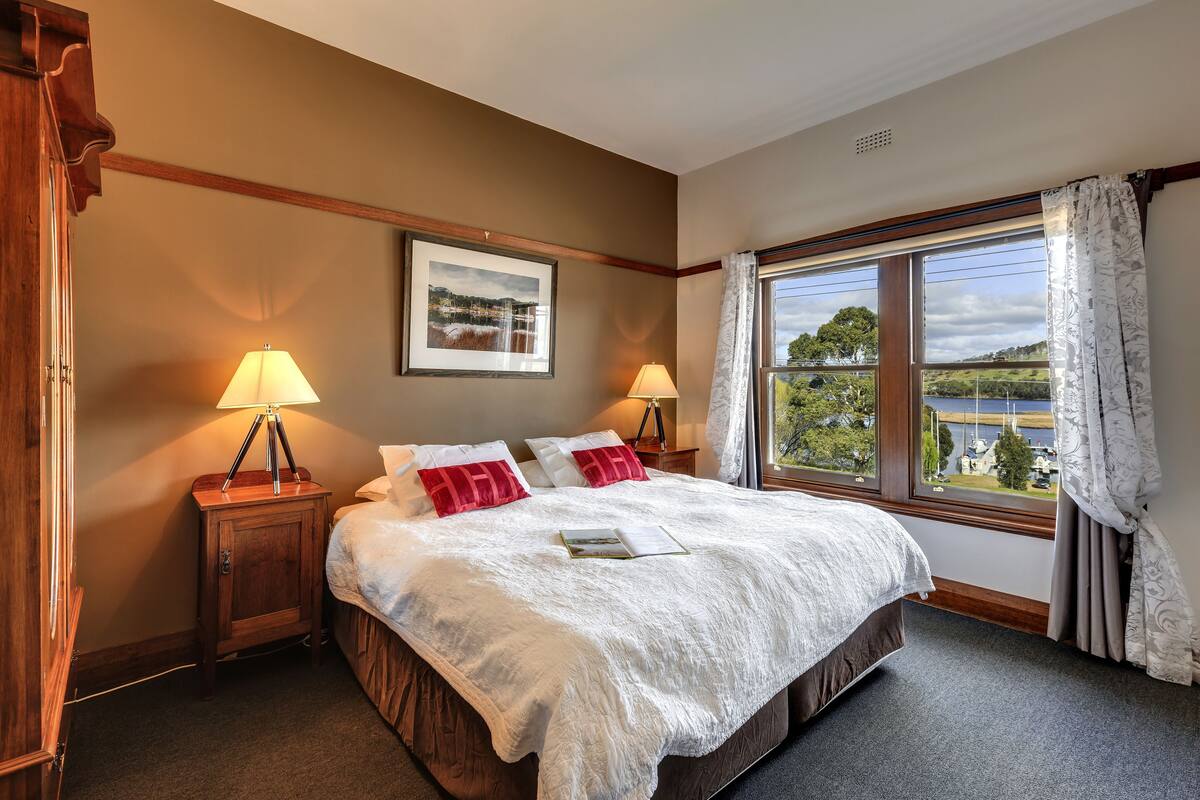 A bedroom with a large bed, two bedside lamps, a framed picture on the wall, and a window with a view of a body of water.