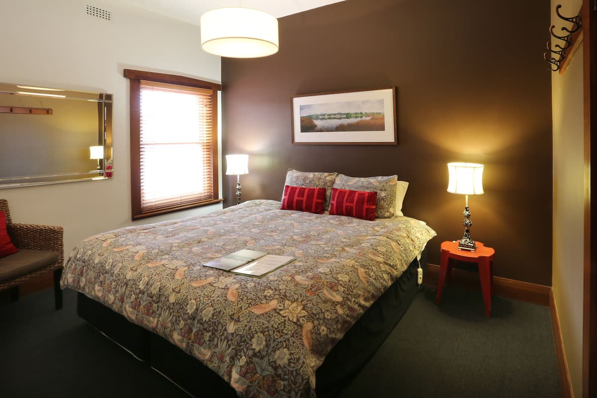 A neatly arranged hotel room with a large bed, a bedside table with a lamp, a chair, and a window with blinds.