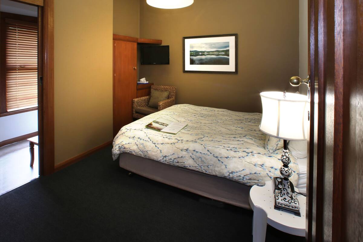 A bedroom with a bed, a chair, a bedside table with a lamp, a framed picture on the wall, and a door with a window.
