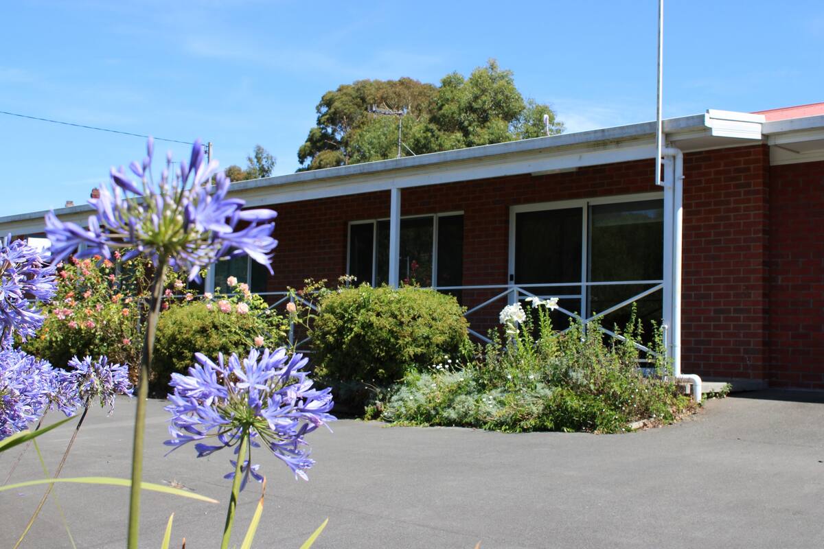 A building with a brick facade, large windows, and a small garden area with purple flowers.
