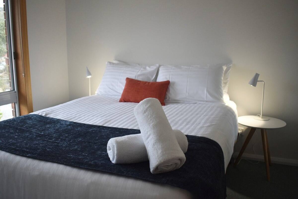 A neatly made bed with white and orange pillows, a dark blue bedspread, and two rolled white towels. There is a small white table with a lamp beside the bed.