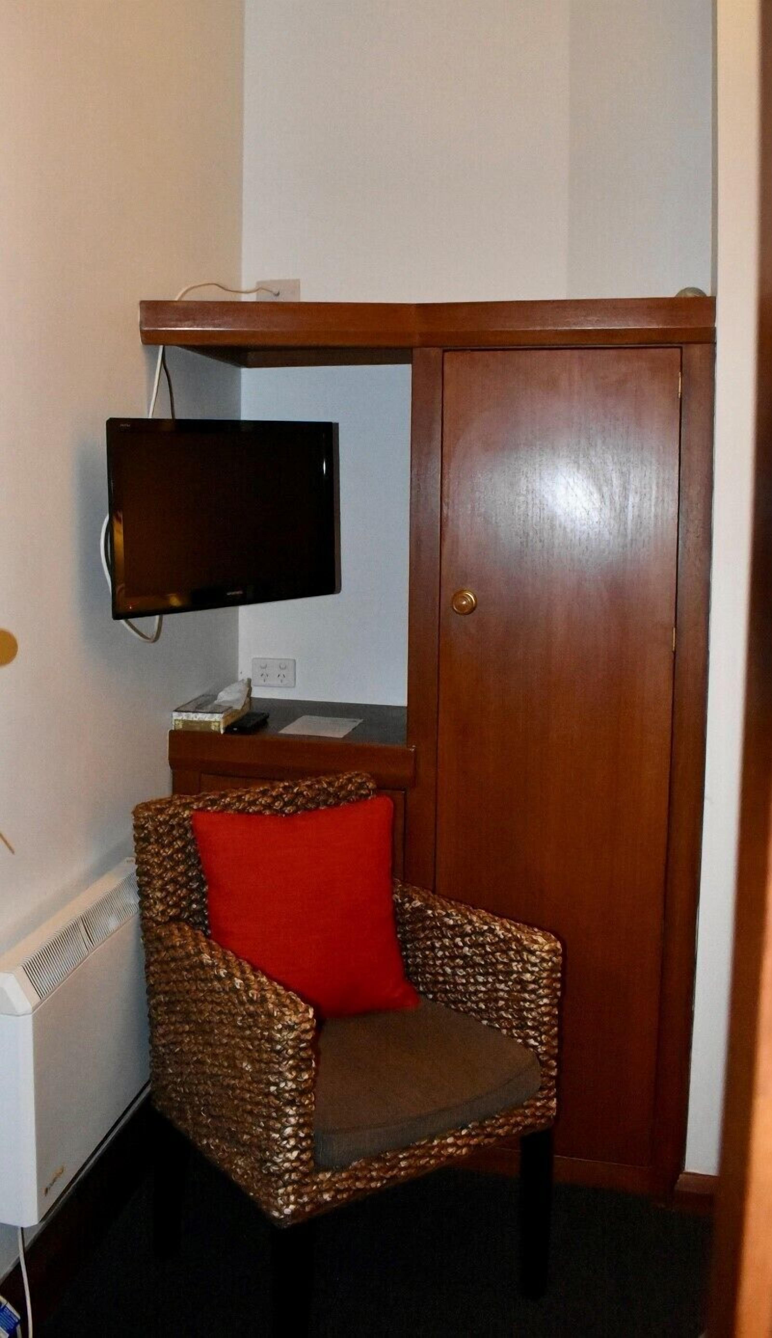 Um quarto pequeno com uma cadeira de vime, uma televisão fixada na parede e um guarda-roupa de madeira.