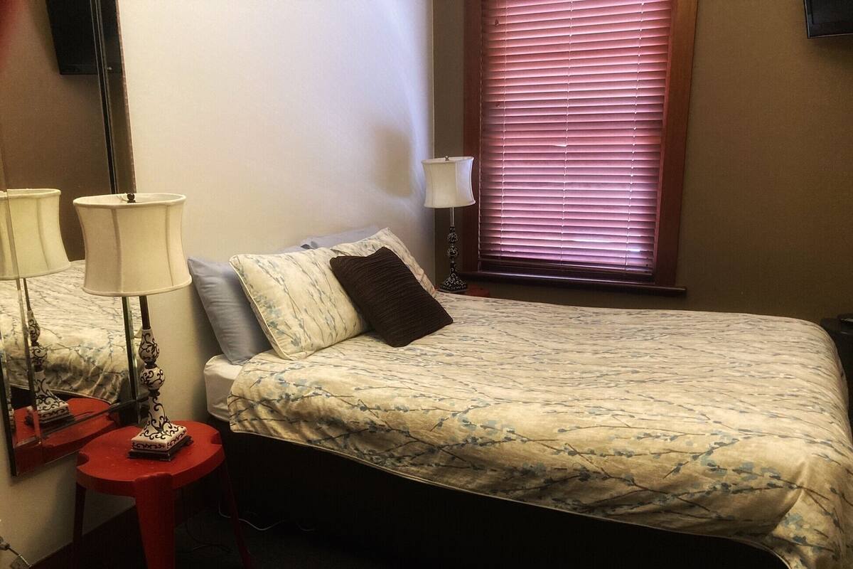 A bedroom with a bed, a nightstand, a lamp, a window with blinds, and a wall-mounted TV.