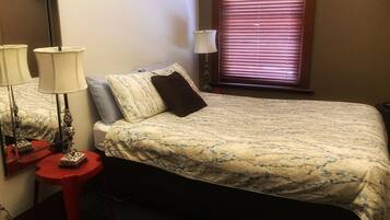 Un dormitorio con una cama, una mesita de noche, una lámpara, una ventana con persianas y un televisor instalado en la pared.