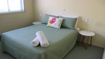 Una cama bien hecha con ropa de cama verde, una almohada estampada con motivos florales y dos toallas blancas. Se observa una mesita de noche con una lámpara blanca.