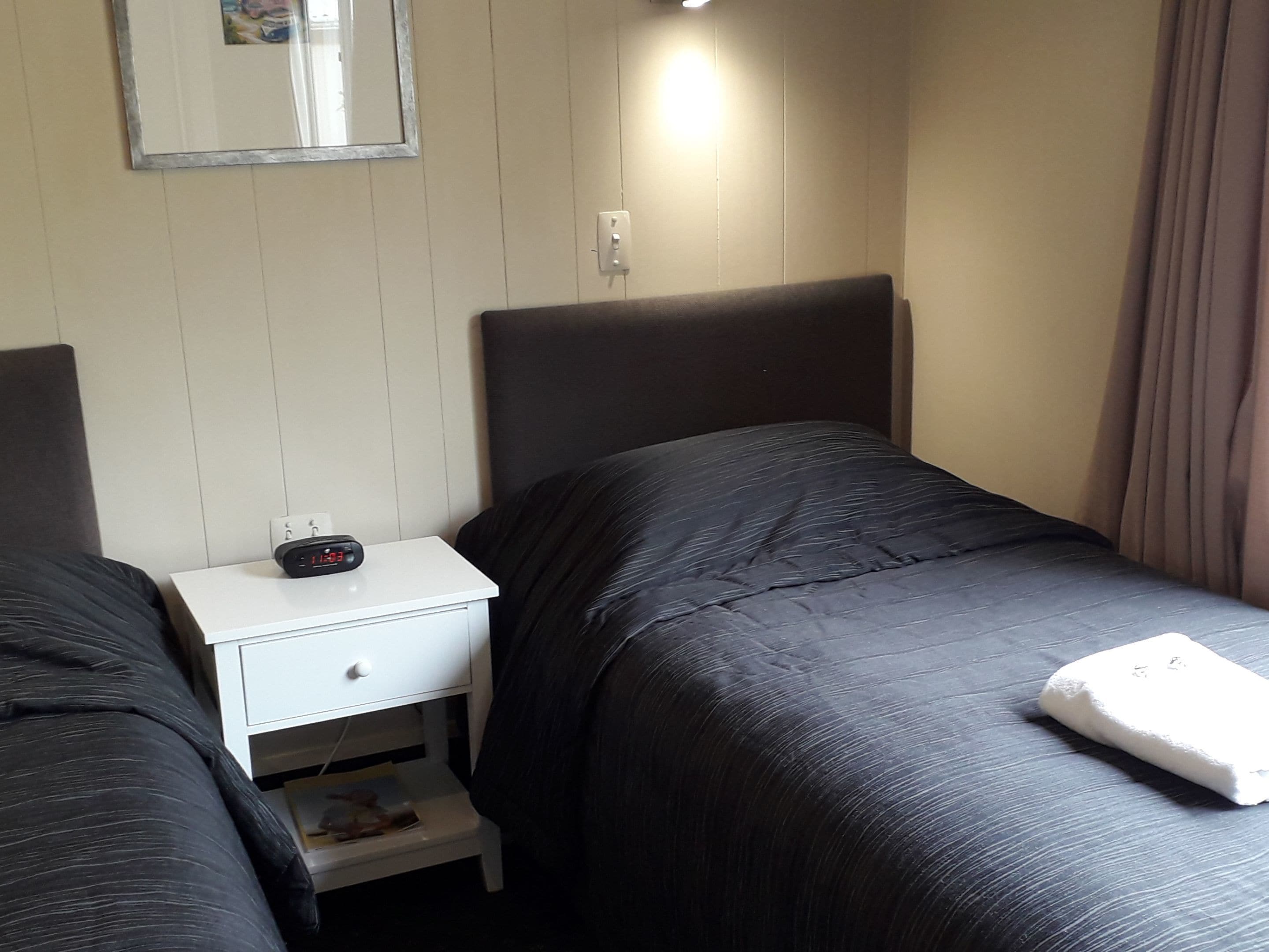 A hotel room with two beds, a nightstand with a clock, a small drawer, and a mirror on the wall.