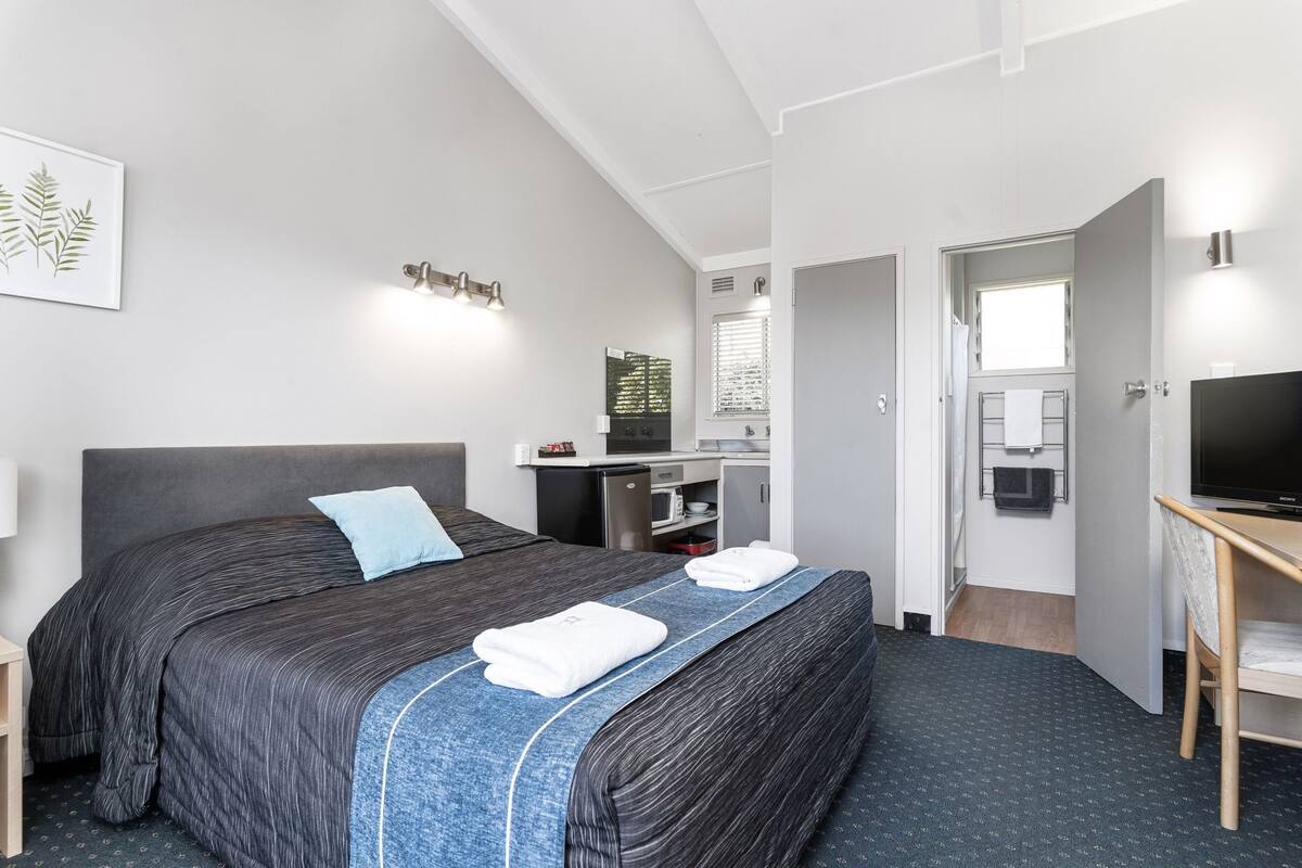 Habitación de hotel moderna con una cama grande, un escritorio con televisor de pantalla plana, una kitchenette compacta y un baño visible a través de una puerta abierta.