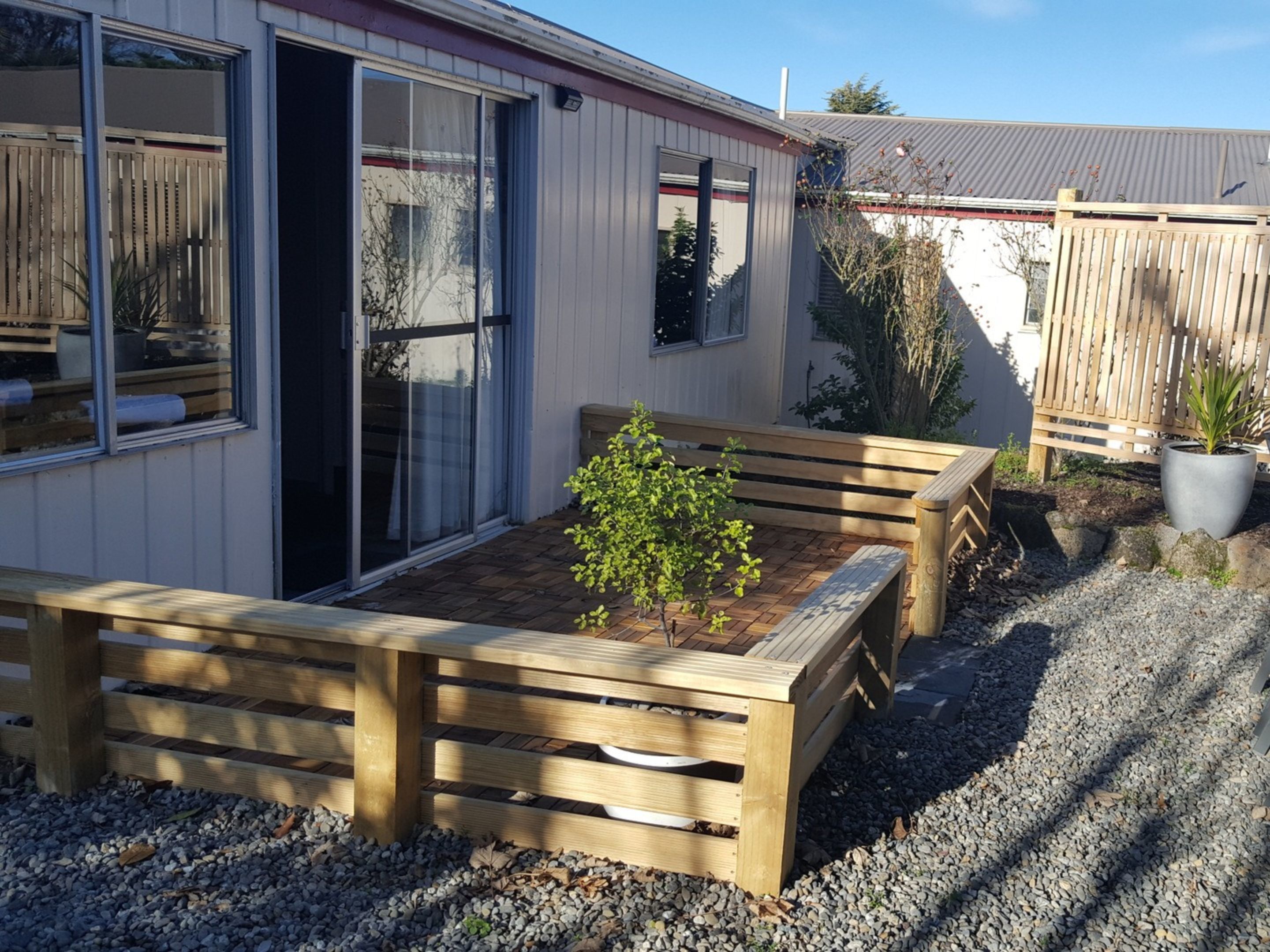 Un banco de madera con un pequeño árbol en un área de grava, una puerta de vidrio corredera y una valla de madera al fondo.