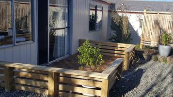 Un banco de madera con un pequeño árbol en un área de grava, una puerta de vidrio corredera y una valla de madera al fondo.