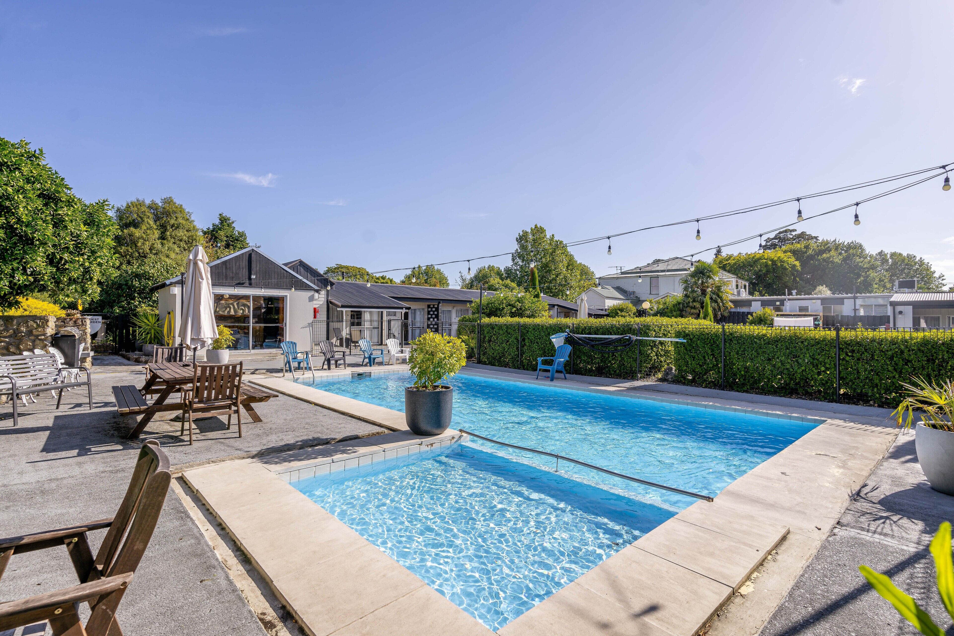A swimming pool with lounge chairs and umbrellas, surrounded by a hedge and a row of houses.