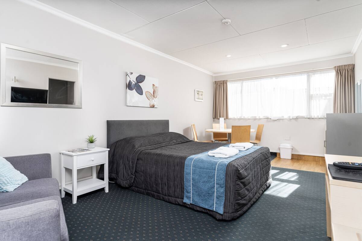 Una habitación de hotel moderna con cama, escritorio, silla, mesita, televisor y ventana con cortinas.