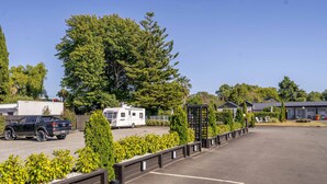 A parking lot with a row of neatly trimmed bushes, a tall evergreen tree, and a row of buildings with a modern design. - Amble Inn Motel - Masterton (Masterton)