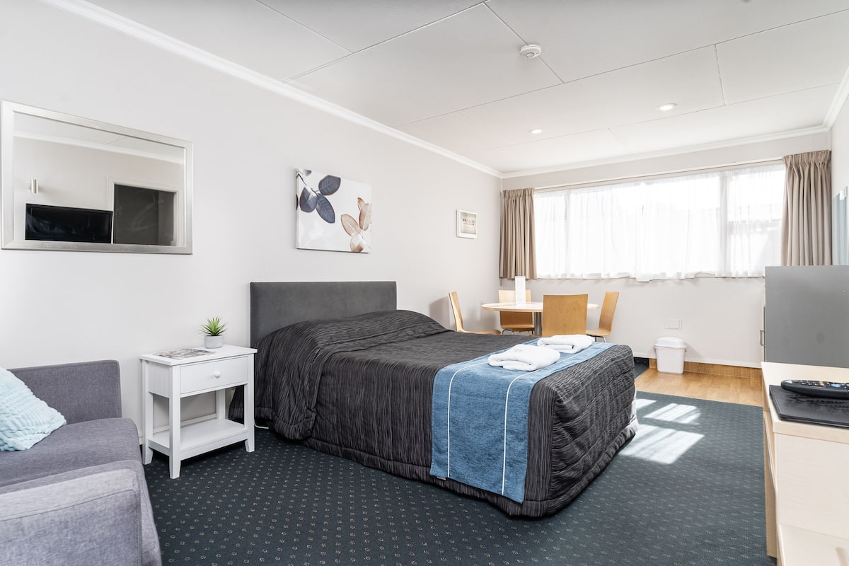 Habitación de hotel con cama, escritorio, silla, una mesita, televisor y ventana con cortinas.