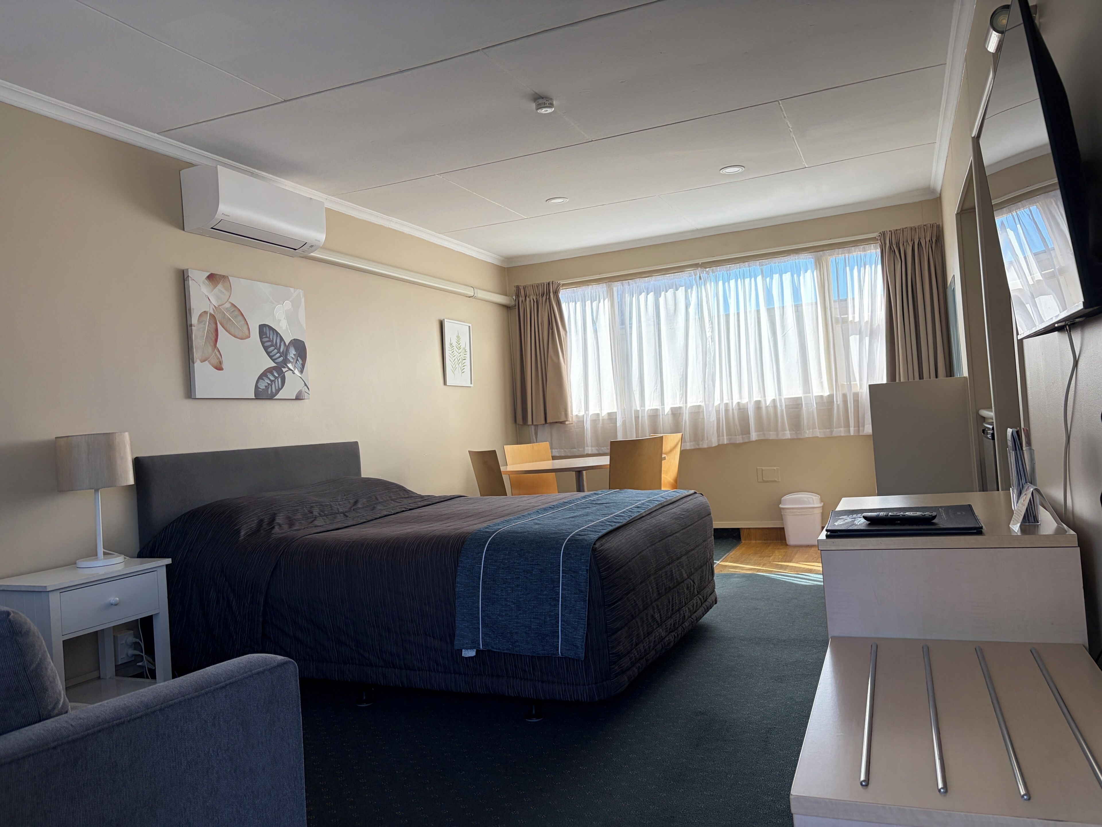 A hotel room with a bed, bedside table, lamp, desk, chair, and a large window with curtains.