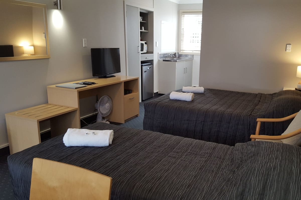 Habitación de hotel con dos camas, un escritorio con televisor, un ventilador y una kitchenette.